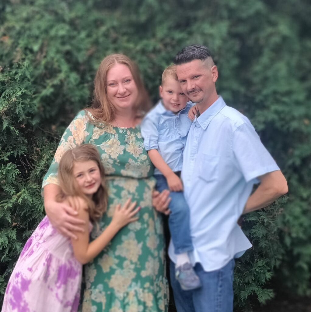 Family portrait of parents with two young children posing together outdoors against green foliage