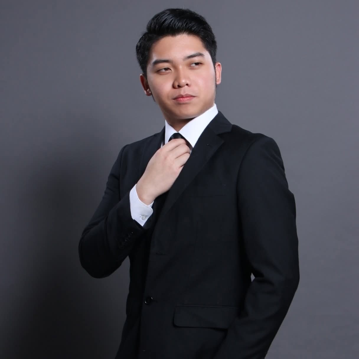 Professional portrait of a young man in a black suit adjusting his tie against a gray backdrop